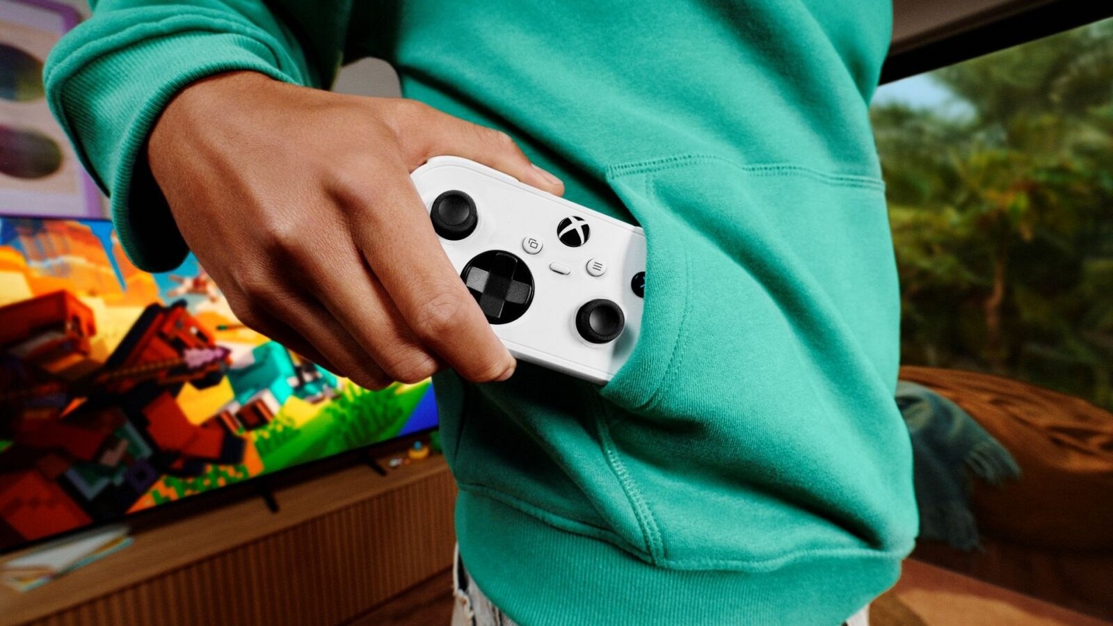 มือถือจอย XBOX Nano Controller สีขาวที่กำลังหยิบออกจากกระเป๋าเสื้อฮู้ดสีเขียว ด้านหลังเป็นทีวีเล่นเกม