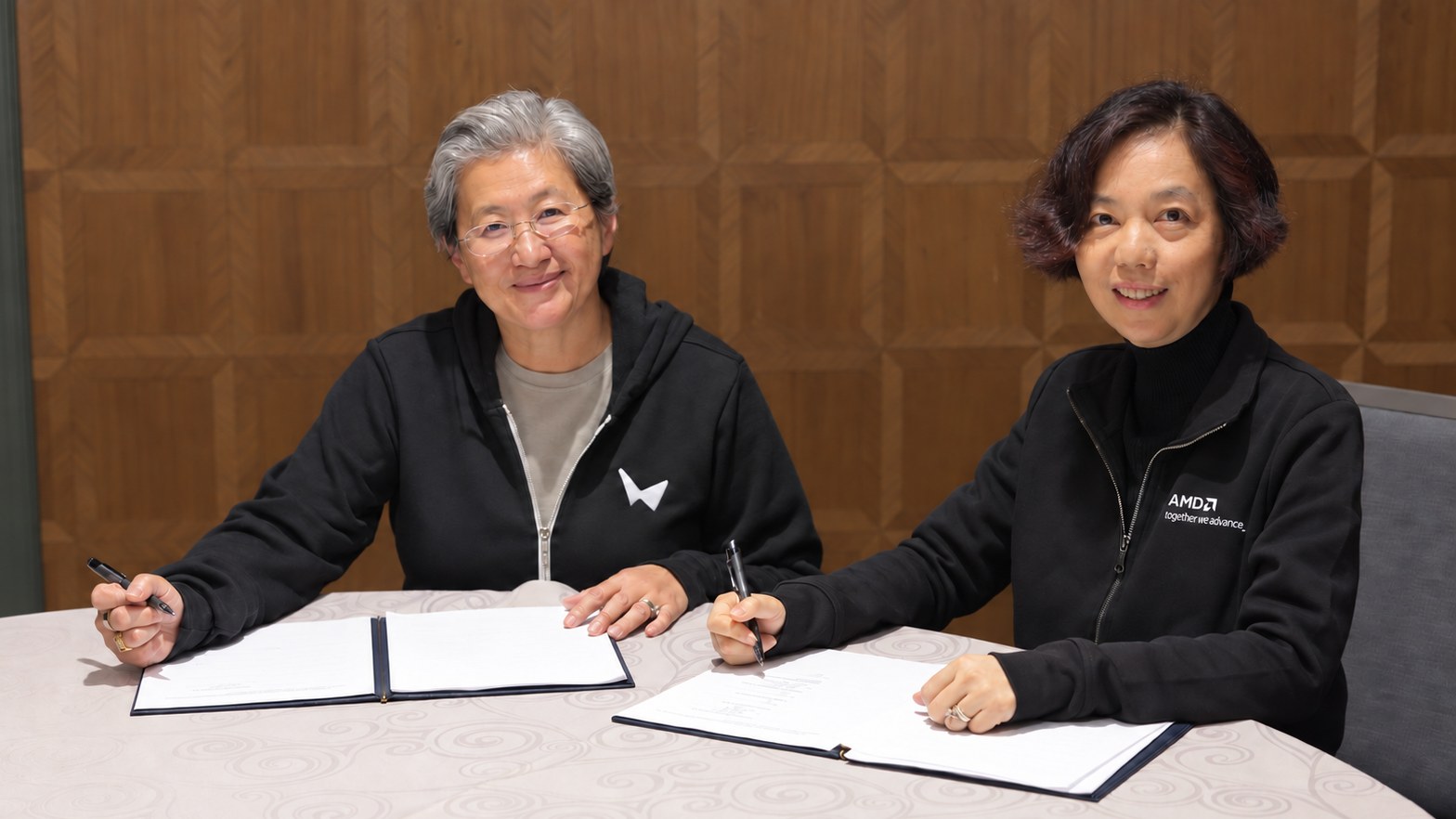 ผู้บริหาร AMD และ World Labs นั่งลงนามเอกสารซื้อกิจการที่โต๊ะ สวมแจ็กเก็ตโลโก้ของสองบริษัท