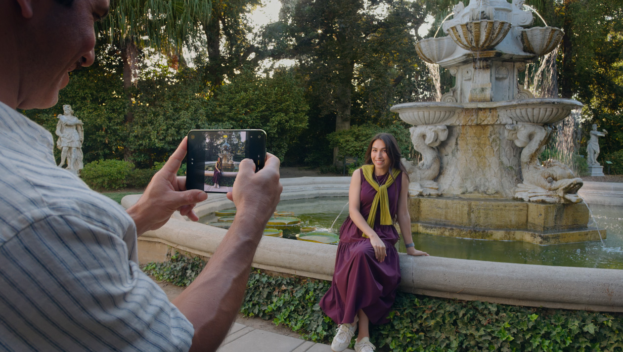 ผู้ใช้ถือ iPhone Duo ถ่ายภาพผู้หญิงที่นั่งข้างน้ำพุ โดยจอด้านนอกของเครื่องแสดงภาพพรีวิวของตัวแบบ